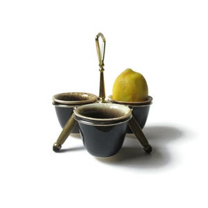 May include: A three-piece ceramic condiment set with a gold metal stand and handle. The set features three small bowls with a black glaze and a white speckled design. A lemon is sitting in one of the bowls.