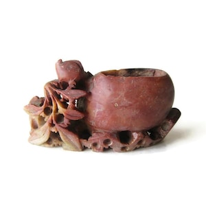 May include: A carved, reddish-brown stone sculpture featuring a central, rounded vessel. The sculpture has intricate floral and leaf details, with a textured surface. The piece is likely a decorative item or a small container.