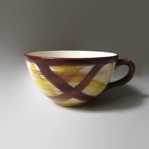 May include: A ceramic teacup with a handle. The cup features a cream-colored interior and a yellow and white exterior with a crisscross pattern in dark brown. The rim and handle are also dark brown.