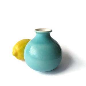May include: A small, round, turquoise ceramic vase with a white interior rim. A yellow lemon is positioned to the left of the vase. The vase has a smooth, glossy finish and a simple, elegant design. The background is white.