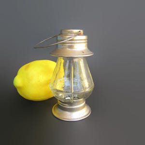 May include: A small, clear glass lantern with a metallic silver-toned top and base, featuring a wire handle at the top. Positioned next to the lantern is a single, bright yellow lemon, providing a clear contrast in color and shape to the glass object.