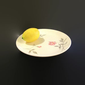 May include: A white ceramic plate with a floral design featuring pink roses and gray leaves. A yellow lemon sits on the plate.