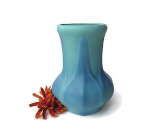May include: A two-toned blue ceramic vase with a bulbous base and a narrow neck. The vase is next to a vibrant orange-red flower, set against a white background. The vase's design is smooth and elegant.
