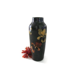 May include: A dark, slender vase with a glossy finish, adorned with hand-painted floral designs in shades of yellow and orange. A small, red flower rests near the base. The vase is approximately 10 inches tall.