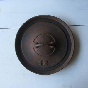 May include: A round, dark brown cast iron lid with a textured, weathered surface. The lid features a central circular handle and the number 12 embossed clearly on the lower portion of the surface. The design includes a raised outer rim edge.