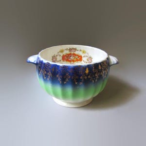 May include: A vintage ceramic bowl with a white interior and a floral design. The exterior features a gradient of green to blue, adorned with gold accents. It has two small handles and a white base.