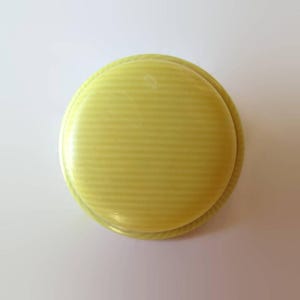May include: A pale yellow, round object with a ridged surface. The object has a slightly raised, rounded edge. The overall shape is reminiscent of a button or a small, decorative element. The color is a soft, muted yellow.