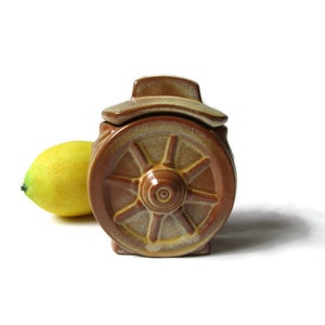May include: A brown ceramic jar with a lid shaped like a wagon wheel. The jar has a light brown glaze with a darker brown rim. The lid is slightly ajar.