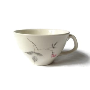 May include: A white ceramic teacup with a delicate floral design featuring a pink rose and gray leaves. The cup has a rounded shape and a handle.