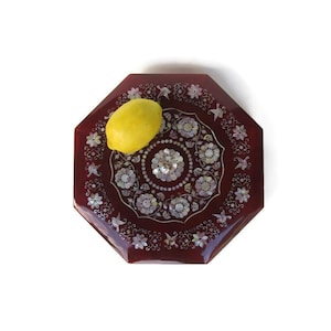 May include: An octagonal, dark red decorative tray with an inlaid floral design and a yellow lemon. The tray's center features a detailed mother-of-pearl floral pattern. The lemon rests on the tray's surface, adding a pop of color.