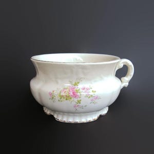 May include: A white porcelain planter with a handle and a scalloped base. The planter is decorated with a floral design of pink roses and green leaves. The rim and base are trimmed with gold.