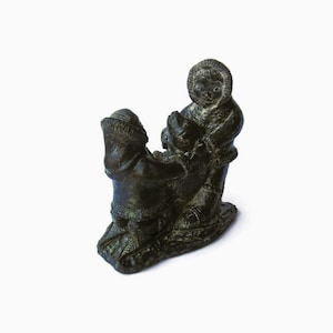 May include: A dark charcoal-colored stone sculpture depicting two figures wearing hooded parkas. One figure is standing while the other is kneeling, both interacting with a central animal form. The piece features textured details on the clothing.