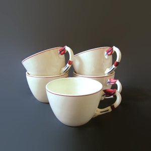 May include: Set of four white ceramic teacups with red and blue painted floral designs on the handles. The cups are stacked on top of each other.
