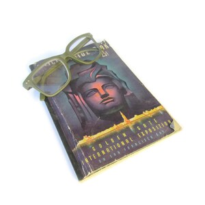 May include: Olive green eyeglasses rest on a vintage book. The book cover features a stylized face and the text "Golden Gate International Exposition on San Francisco Bay". The book is slightly worn.
