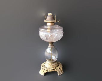 May include: An antique oil lamp with a clear, cut-glass reservoir and globe. The lamp features a brass base and accents, with a decorative, ornate design. The lamp is designed to sit on a table or other flat surface.