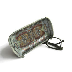 May include: A rectangular, curved metal tray featuring a metallic finish with intricate, multi-colored geometric and scrollwork patterns in shades of copper, silver, and gold, paired with a pair of black-framed eyeglasses resting on the surface.
