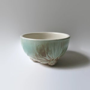 May include: A small, light green ceramic bowl with a textured, leafy design. The bowl has three small, rounded feet.