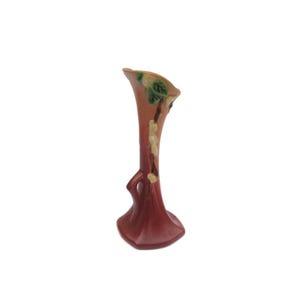 May include: A small, antique vase with a flared top and a dark red base. The vase has a unique shape with a cutout near the base and is decorated with green and white floral accents. The vase is approximately 7 inches tall.