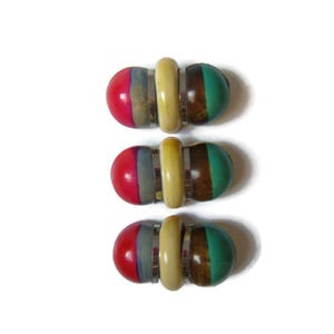 May include: Three decorative barrettes with a unique design. Each barrette features a central, light-yellow cylindrical piece flanked by two rounded, colorful sections. The sections are red, green, brown, and gray, with a silver-colored metal band.