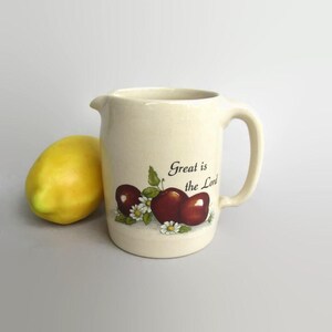 May include: A cream-colored ceramic pitcher featuring a painted illustration of two red apples, green leaves, and white daisies. The text "Great is the Lord" is printed in black script above the fruit. A bright yellow lemon sits to the left.