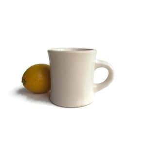 May include: A white ceramic mug with a rounded handle. The mug has a slightly flared rim and a smooth, matte finish.