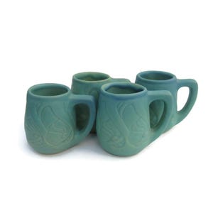 May include: A set of four matching mugs in a matte seafoam green finish. Each mug features a rounded body with a thick, sturdy handle and a raised, embossed floral pattern wrapping around the exterior surface of the ceramic drinkware.