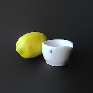 May include: A bright yellow lemon next to a small, white ceramic bowl with a pouring spout. The bowl has blue text that reads "COORS U.S.A.". The objects are set against a dark background.