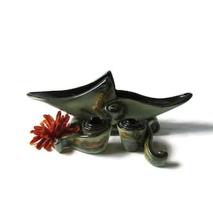 May include: A green and brown ceramic planter with a unique, abstract design. The planter is shaped like a boat with a long, pointed bow and a wide, flat stern. It has three scroll-like legs and a decorative floral pattern.