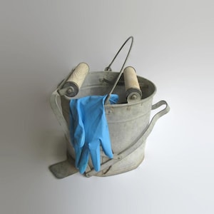 May include: A galvanized metal bucket featuring a wringer mechanism with two wooden rollers and a metal handle. A single light blue rubber glove is draped over the front of the bucket, which has a wide rim and a sturdy base for stability.