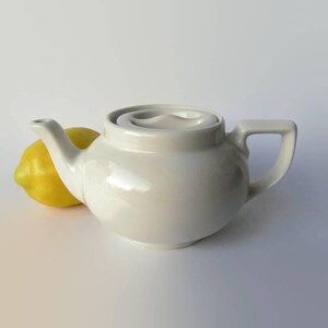 May include: A white ceramic teapot with a rounded body, a spout, and a square handle. A yellow lemon rests beside the teapot. The teapot has a unique swirl design on the lid. The teapot is a kitchenware item.