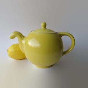 May include: A yellow ceramic teapot with a rounded body, curved spout, and a loop handle. The lid has a small knob on top. A yellow lemon is next to the teapot. The teapot is approximately 8 inches wide.