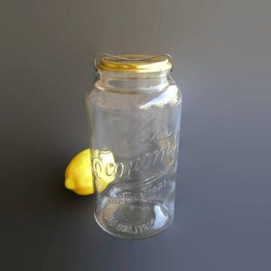 May include: A clear glass jar with a gold-colored metal lid and a wire bail closure. The glass features embossed text that reads Kerr Economy. A single yellow lemon is positioned behind the jar, partially visible through the clear glass walls.