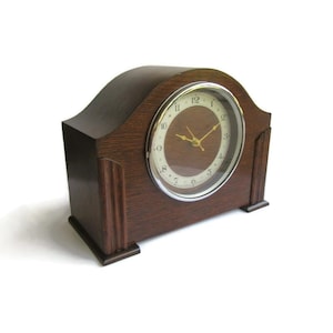 May include: A dark brown wooden mantel clock with a curved top and vertical fluted accents on the sides. The clock face is circular with a cream-colored background, black Arabic numerals, and thin gold-colored hour, minute, and second hands.