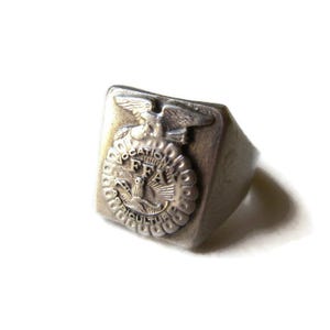 May include: A silver-toned ring with a rectangular face. The face features a detailed emblem with an eagle, the letters "FFA", and the words "Vocational Agriculture". The ring has a slightly worn appearance.
