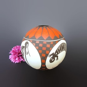 May include: A round, hand-painted ceramic vase with a vibrant orange top and geometric patterns. The vase features black and white illustrations of figures and a lizard. A pink flower rests beside the vase.