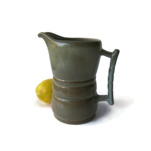 May include: A sage green ceramic pitcher with a glossy finish, featuring a wide spout, a rounded body with two horizontal ridges, and a textured, segmented handle. A single yellow lemon is positioned behind the base of the pitcher.