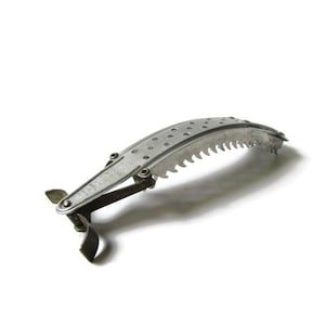 May include: A vintage, silver-colored, hand-operated tool with a curved, serrated blade. The tool has a handle and a spring mechanism. The design includes small holes along the blade's top edge. The tool is likely used for cutting or trimming.