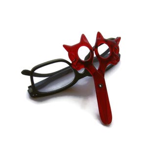 May include: A pair of black-framed eyeglasses with a red cat-shaped scissor-like accessory. The accessory is made of a translucent red material and features cat head designs at the top of each scissor blade.