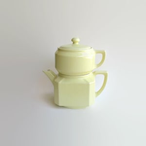 May include: A pale yellow ceramic teapot for one, featuring a stackable design with a matching lid. The base has an octagonal shape with a handle and a spout, while the top section is cylindrical with a handle and a small knob on the lid.