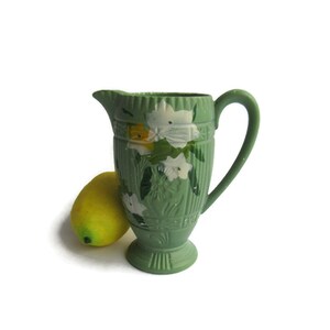 May include: A green ceramic pitcher with a floral design. The pitcher has a white and yellow floral pattern on a green background. The pitcher has a handle and a base.