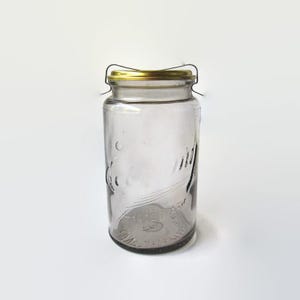 May include: A clear glass jar with embossed lettering on the front surface. The jar features a gold-colored metal lid secured by a thin wire bail closure mechanism that arches over the top. The glass body is cylindrical with a wide mouth opening.