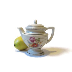 May include: A small white teapot with a floral design and gold accents. The teapot has a lid and a spout. It is sitting on a white surface.