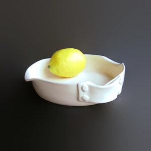 May include: A white ceramic serving dish with a rounded shape and a handle on each side. The dish has a textured surface and is decorated with small metal rivets. A yellow lemon sits in the center of the dish.