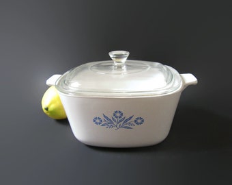 May include: White square baking dish with a glass lid and blue floral design. The dish is on a gray surface.