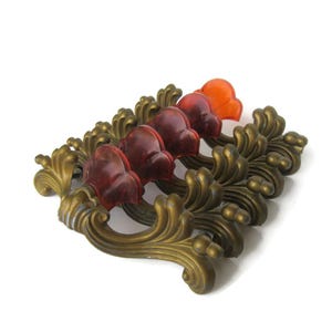 May include: Antique brass-toned drawer pulls with red and orange flower-shaped accents. The decorative hardware features ornate, scrolling details and a vintage aesthetic. The pulls are likely made of metal and plastic.