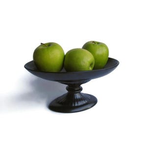 May include: A black, pedestal fruit bowl holds three green apples. The bowl has a wide, shallow dish and a decorative, fluted base. The apples are a vibrant green color.