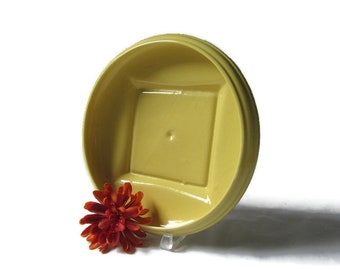 May include: A yellow ceramic dish with a square inset in the center. The dish is on a white surface with a red flower in the foreground.