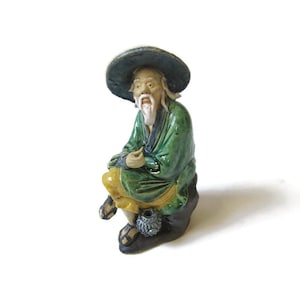 May include: A ceramic figurine of an elderly man wearing a wide-brimmed hat and green robe, seated on a dark rock. The figure has a long white beard and is holding a small object. The figurine has yellow pants and sandals.