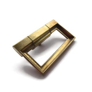 May include: A rectangular brass-colored metal buckle with a hinged section and a rectangular opening. The buckle has a slightly worn appearance, suggesting it is vintage or has been used. The design is simple and functional.