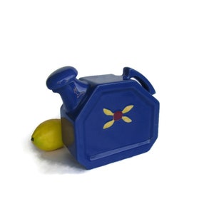 May include: A blue ceramic pitcher with a yellow and red design. The pitcher has a spout and a handle. It is sitting on a white surface.